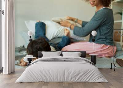 Cropped shot of cheerful mixed race young female smiling, sitting on bed with little son lying with his face down after he jumped off stack of pillows. Fun, entertainment, home and social distancing Wall mural