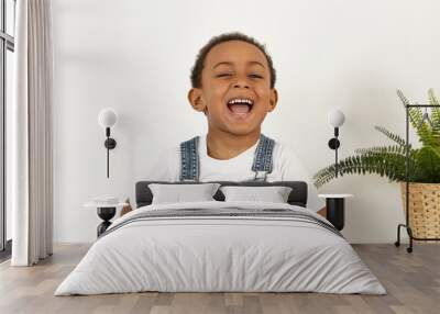 Cute emotional dark skinned little boy posing at white wall with plant in background, opening mouth widely, laughing at funny joke or watching cartoon. Positive emotions, reaction and feelings Wall mural