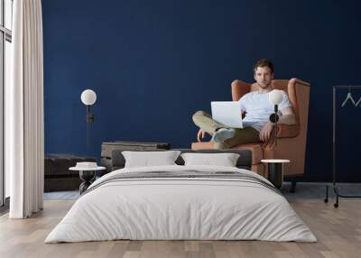 Fashionable young Caucasian man with beard relaxing in modern loft space interior, sitting on orange armchair and surfing internet on laptop pc. Handsome guy enjoying online communication using gadget Wall mural