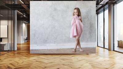 Full length portrait of beautiful barefooted timid European little girl with long loose hair standing on wooden floor at home, crossing her legs, feeling shy while posing in new pink fancy dress Wall mural