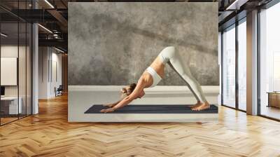 Full length side view of beautiful young fit woman in sportswear working out indoors, practicing yoga exercise on mat, doing downward facing dog pose or Adho Mukha Svanasana sun salutation pose Wall mural