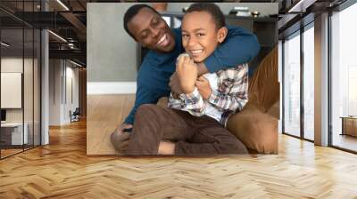 Funny picture of caring and loving dark-skinned dad hugging his misbehaving, naughty boy, both having fun, laughing, spending leisure time enjoying bonding and warm relationships, sitting on floor Wall mural