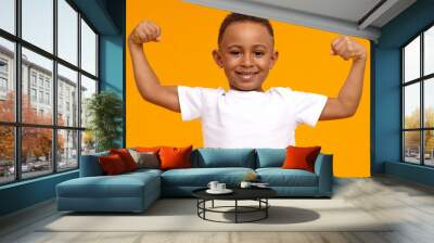 Handsome confident Afro American eight year old child in casual t-shirt smiling happily and raising clenched fists, tensing muscles, feeling strong and full of energy after ate healthy protein lunch Wall mural