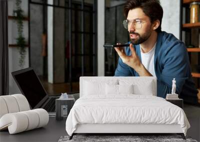 Handsome man at office sitting at working table with laptop with copy space screen for your advertising content recording voice mail or using virtual assistant to find information in internet Wall mural