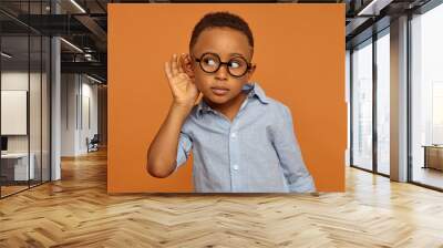 I want to hear everything. Handsome nosy African American little boy wearing round spectacles and neat shirt having curious facial expression while spying, overhearing, holding hand on his ear Wall mural