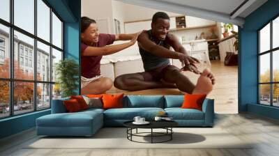 Indoor image of beautiful young Latin female laughing while sitting on floor pushing her funny inflexible black boyfriend who is doing forward bend. People, relationships and active lifestyle Wall mural