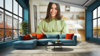 Indoor image of stylish young 20 year old brunette woman dressed in trendy hooded sweatshirt standing in cozy home interior, keeping arms on her chest and smiling after running workout outdoors Wall mural