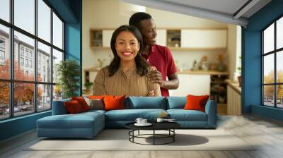 Indoor picture of pretty charming lady of 30s in brown casual dress standing with arms crossed and adorable smile looking at camera while her ethnicity boyfriend in red hugging her shoulders Wall mural