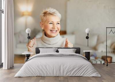 Indoor shot of fashionable overjoyed mature female in turtleneck sweater enjoying positive news, having ecstatic facial expression, laughing and clenching fists. Success and achievements concept Wall mural