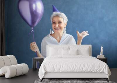 Joy, happiness, fun and positive emotions concept. Studio shot of beautiful excited 50 year old gray haired woman with conical hat on her head, celebrating anniversary, gesturing emotionally Wall mural