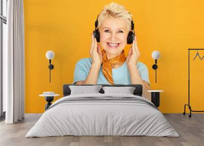 Modern stylish Caucasian woman pensioner with short hairstyle relaxing listening to favorite tracks via earbuds. Attractive mature female enjoying nice music using wireless Bluetooth headphones Wall mural