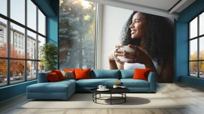 Morning routine. Portrait of happy charming young mixed race female with wavy hair enjoying summer view through window, drinking good coffee, sitting on windowsill and smiling. Beautiful daydreamer Wall mural