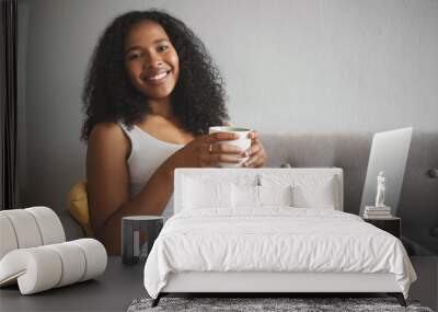 People, leisure, technology and gadgets concept. Portrait of stylish beautiful young African American woman with wavy hair enjoying slow morning, having cup of coffee and surfing internet using laptop Wall mural