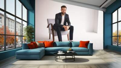 People, lifestyle, business, style, fashion and men's wear concept. Positive successful young CEO sitting in armchair, smiling at camera, dressed in elegant shoes, trousers, jacket and white t-shirt Wall mural