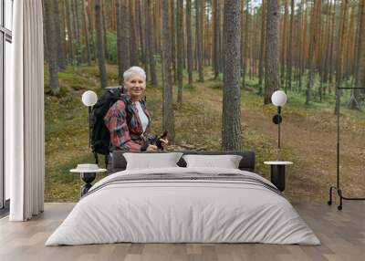 Portrait of attractive middle aged female in activwear standing on trail in national park using poles for nordic walking, enjoying nice fresh air among pine trees, looking at camera with smile Wall mural