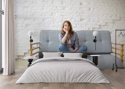 Portrait of beautiful pensive young Caucasian female keeping legs crossed while sitting on couch against white brick wall background, having thoughtful or bored look, dreaming of something pleasant Wall mural