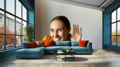 Portrait of cheerful laughing girl of 20s in yellow sweater putting hand next to ear listening to funny conversation, isolated on gray studio background with copy space on the left. Curiosity concept Wall mural