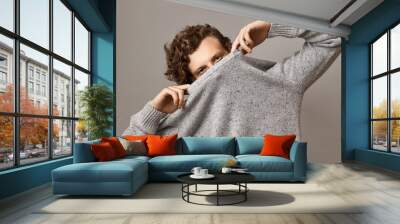 Portrait of trendy looking cute young Caucasian male with wavy voluminous curly hairstyle fooling around, pulling gray sweater over his face, leaving eyes open, having confident facial expression Wall mural