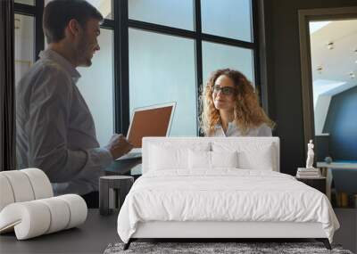 Portrait of two professionals male and female standing by the window opposite each other, smiling while discussing business details, plans and strategy, using laptop computer and making notes Wall mural