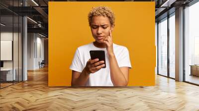 Puzzled focused concerned black female trying to use or download new banking application and making transactions online standing against yellow background, frowning and touching her cheek thinking Wall mural