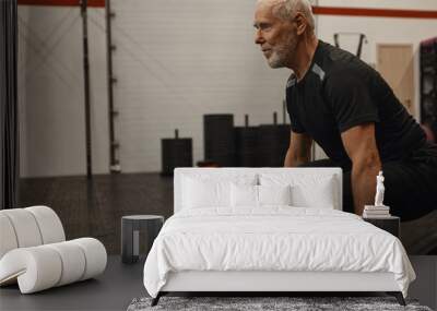 Side view of confident senior male bodybuilder with gray beard doing squats indoors holding barbell. Attractive muscular retired man exercising at fitness center, shaping up, gaining srong body Wall mural