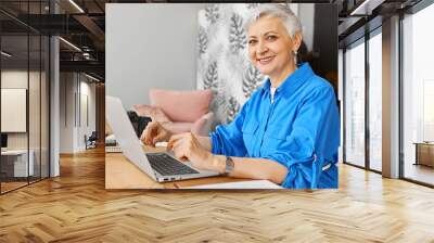 Side view of successful mature female blogger sitting at home office with digital camera and open laptop, keyboarding, writing new article on psychology, looking at camera with broad confident smile Wall mural