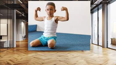 Strength, healthy lifestyle, activity, vitality and sports concept. Indoor shot of serious confident energetic dark skinned athletic boy tensing arm muscles, showing biceps, sitting on mat Wall mural