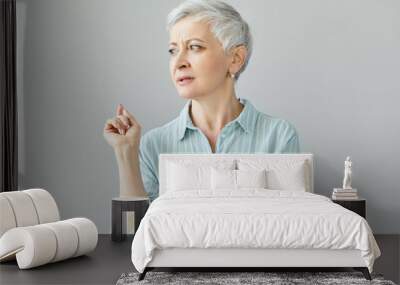 Stylish mature woman professor or couch with pixie haircut looking away, raising fore finger, saying something, giving lecture, sharing her life experience. Studio shot of mature female telling story Wall mural