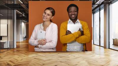 Stylish young mixed race team of two coworkers using electronic gadgets for work, positive cheerful white female carrying digital tablet and handsome smiling black male embracing laptop computer Wall mural