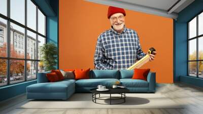 Successful professional attractive elderly male construction worker wearing overalls and belt kit of instruments posing in studio, smiling happily, measuring length of wooden plank using tool Wall mural