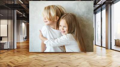 Two happy kids standing at blank grey wall and embracing. Adorable pretty little girl with long hair hugging tight cute blonde boy, showing her love and care. Brother and sister having fun at home Wall mural