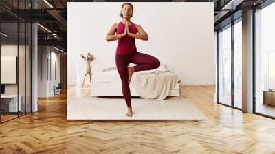 Well being, active lifestyle, health and fitness concept. Portrait of healthy young mixed race woman with strong flexible body, standing upright on carpet, doing tree pose, training balance Wall mural