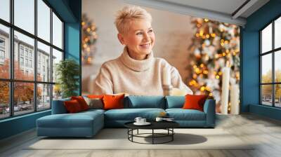 Xmas, holidays, decor, party and festive atmosphere concept. Good looking cheerful middle aged female with short hair enjoying Christmas mood, sitting around wax candles, decorations and lights Wall mural