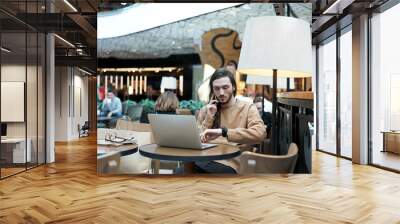 Young business man waiting for a meeting in cafe in shopping center, Long hair guy talk bu phone and watch time. Student making freelance job out of home with laptop and wifi connection. Wall mural