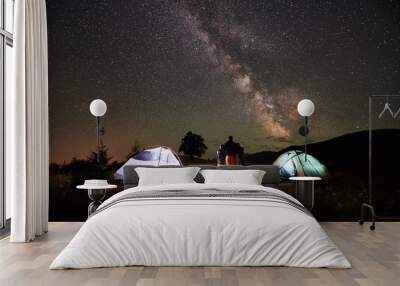 Back view of mother and two sons backpackers resting at camping in mountains, standing beside campfire and two illuminated tents, looking at night sky full of stars, Milky way, enjoying night scene. Wall mural