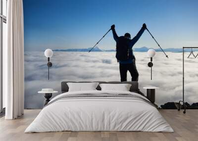 Back view of tourist hiker with backpack standing on top of rocky mountain with trekking poles in raised arms on blue sky at dawn, foggy valley filled with white clouds and mountain tops background. Wall mural