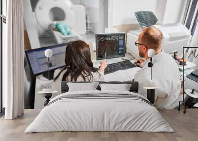 Back view of two doctors sitting, working at computer, examining MRI results. Specialists wearing white robes, consulting, woman pointing by pen, patient lying. Concept of medicine. Wall mural