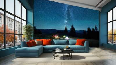 Blurred silhouettes of tourists resting at campsite around campfire with glowing tents nearby under starry sky. Milky Way stretches above, casting magical light over tranquil mountain landscape. Wall mural