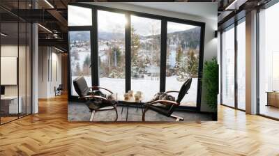 Cozy room with small coffee table and armchairs in front of large panoramic window. Front view of two wooden chairs and table against wide window with snowy mountain landscape. Concept of holidays. Wall mural