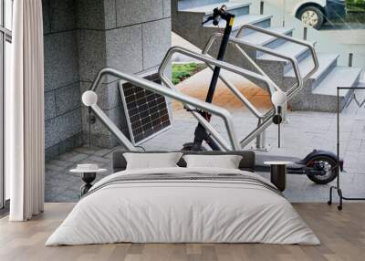 Electric scooter parked in modern urban setting next to solar panel positioned on ground. Sleek gray stone walls and stairs, with metal railings. Sustainable energy solution for urban commuting. Wall mural