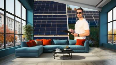 Handsome stylish guy in sunglasses makes an entry in folder against the background of rows solar panels under blue sky. Man with watch in white T-shirt and jeans is smiling while looking at camera Wall mural