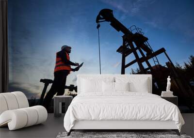 Low angle side view snapshot of petroleum engineer wearing orange vest and a helmet, looking at the oil rig and making some notes against beautiful evening sky, sunset Wall mural