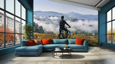 Man riding bicycle on grassy hill and looking at beautiful misty mountains. Male bicyclist enjoying panoramic view of majestic mountains during bicycle ride. Concept of sport, bicycling and nature. Wall mural