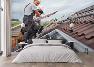 Men technicians carrying photovoltaic solar moduls on roof of house. Builders in helmets installing solar panel system outdoors. Concept of alternative and renewable energy. Wall mural