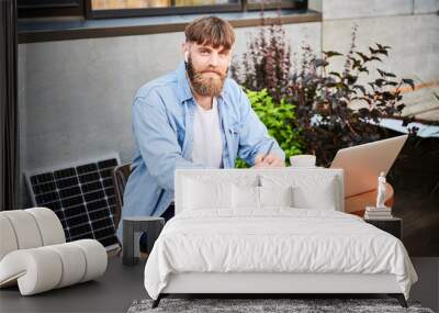 Portrait of bearded man works at round wooden table with laptop, notebook, and cup of coffee. Guy wears earbuds, surrounded by greenery and solar panel, highlighting modern, productive workspace. Wall mural