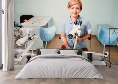 Portrait of woman gynecologist working in gynecological cabinet with modern equipment. Doctor sitting in front of gynecological chair and holding colposcope. Concept if gynecology and medical workers. Wall mural