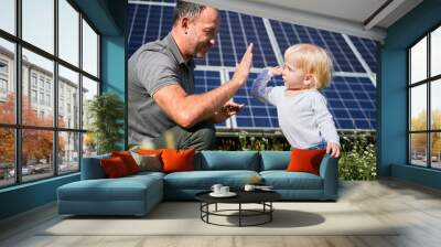 Smiling father and his little child giving each other high-five on background of solar panels. Young father enjoy spending time with his son. Happy family of two on background of solar panels. Wall mural