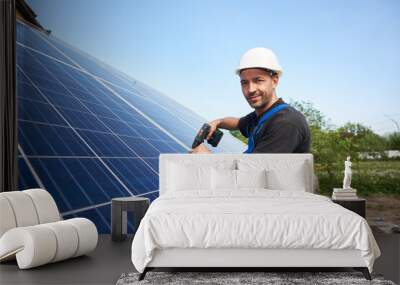 Smiling technician mounting solar panel to metal platform using electrical screwdriver on blue sky copy space background. Stand-alone solar system installation, efficiency concept. Wall mural