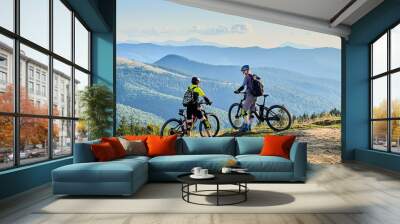 Two cyclists men riding electric bikes outdoors. Back view of male tourists resting on the top of hill, enjoying beautiful mountain landscape, wearing helmet and backpack. Concept of active leisure. Wall mural