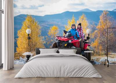 Two female riders ATV in jackets and hats on a snow-covered trail at sunny autumn day against trees with yellow leaves and mountains. Blurred background Wall mural